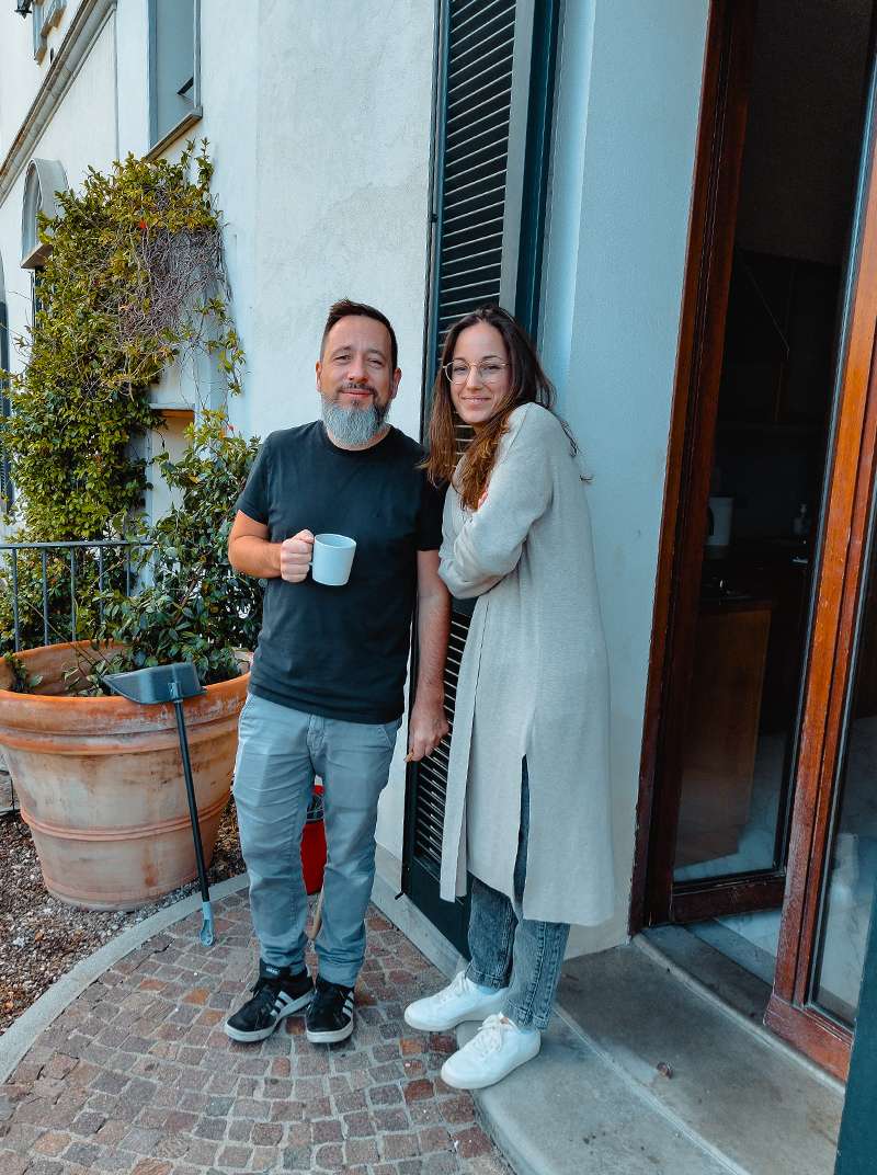 Arne, der eine Kaffeetasse hält und Sarah, die die Arme vor dem Bauch verschränkt hat, stehen vor dem Haus und machen Pause.