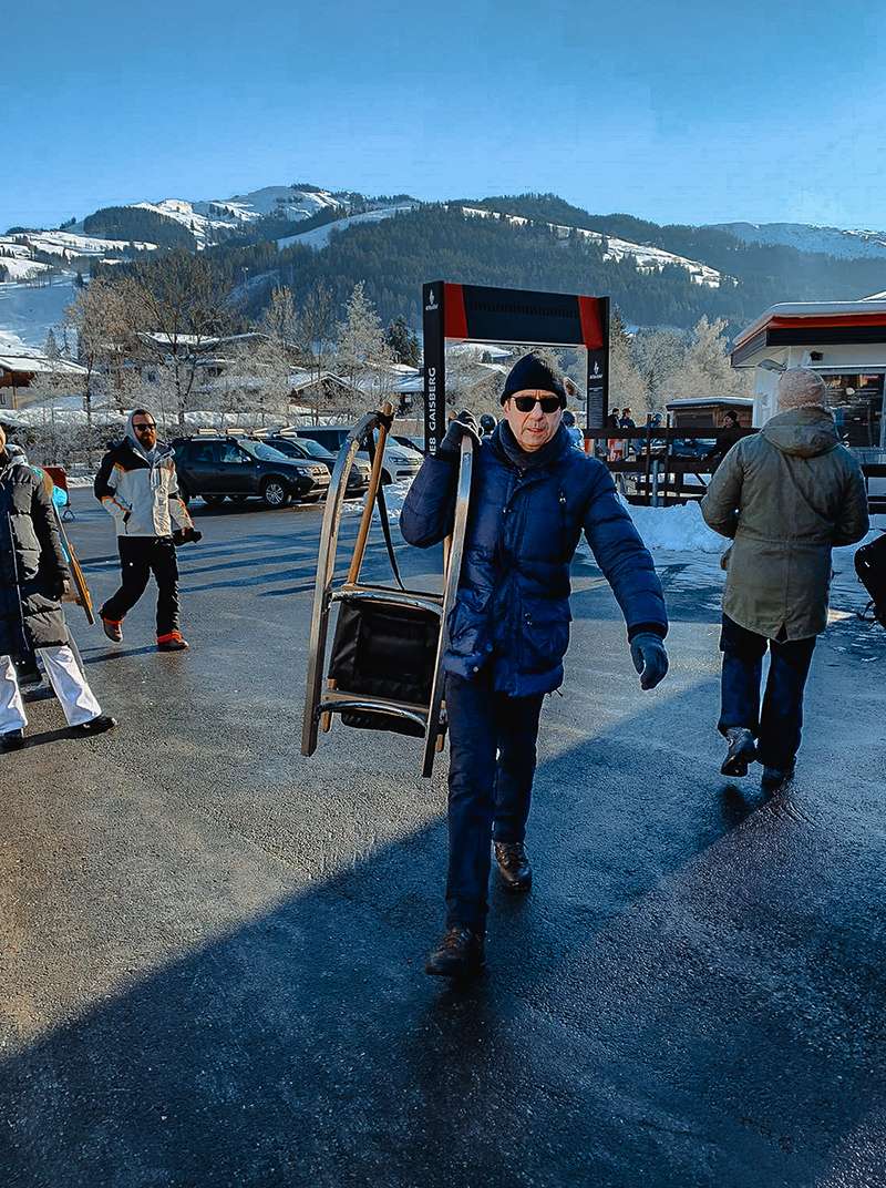 Lothar steht in Winterkleidung mit einem Schlitten über der Schulter auf einem Parkplatz vor einer Bergkulisse.