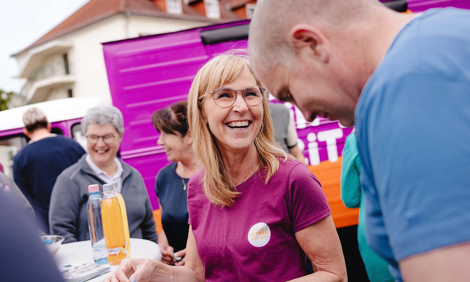 Mehrere lachende Menschen stehen um einen Stehtisch herum. Die Frau in der Bildmitte trägt ein violettes T-Shirt mit der Aufschrift “Viele Extras. Viel Wir.”