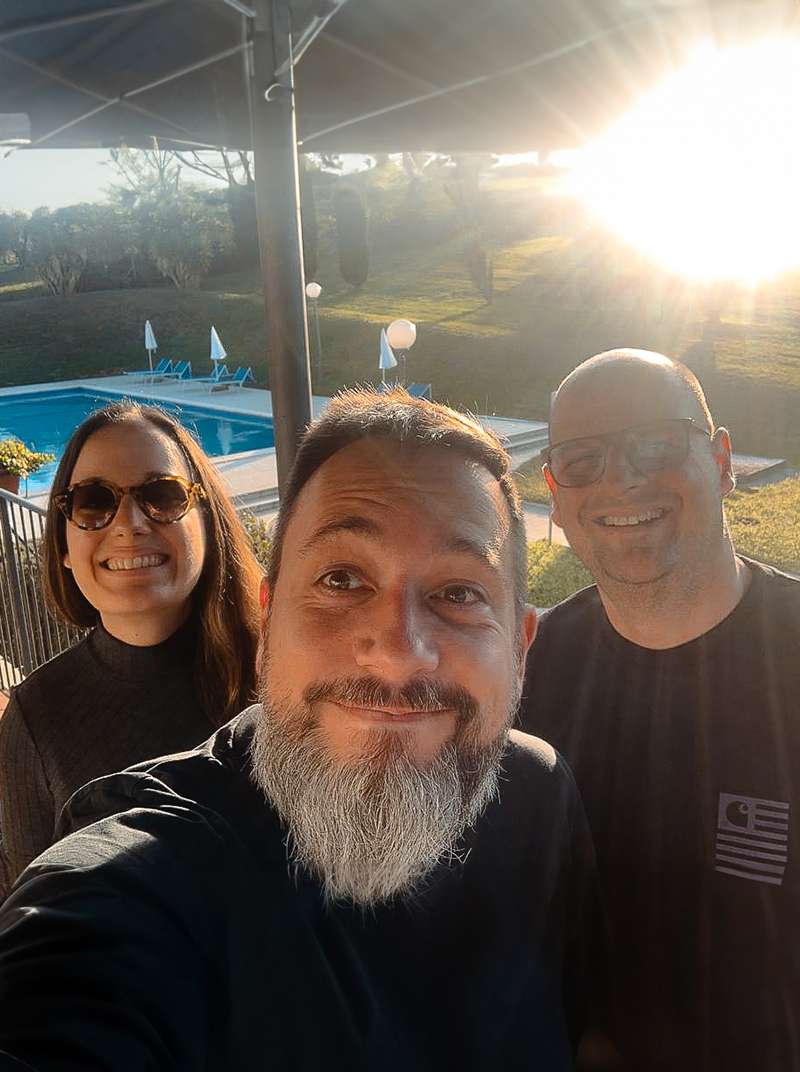 Selfie von Sarah, Arne und Roland vor dem Pool in Italien auf der Workation. Alle lachen und die Sonne scheint hinter Roland, so dass sich Lichtreflexionen bilden.